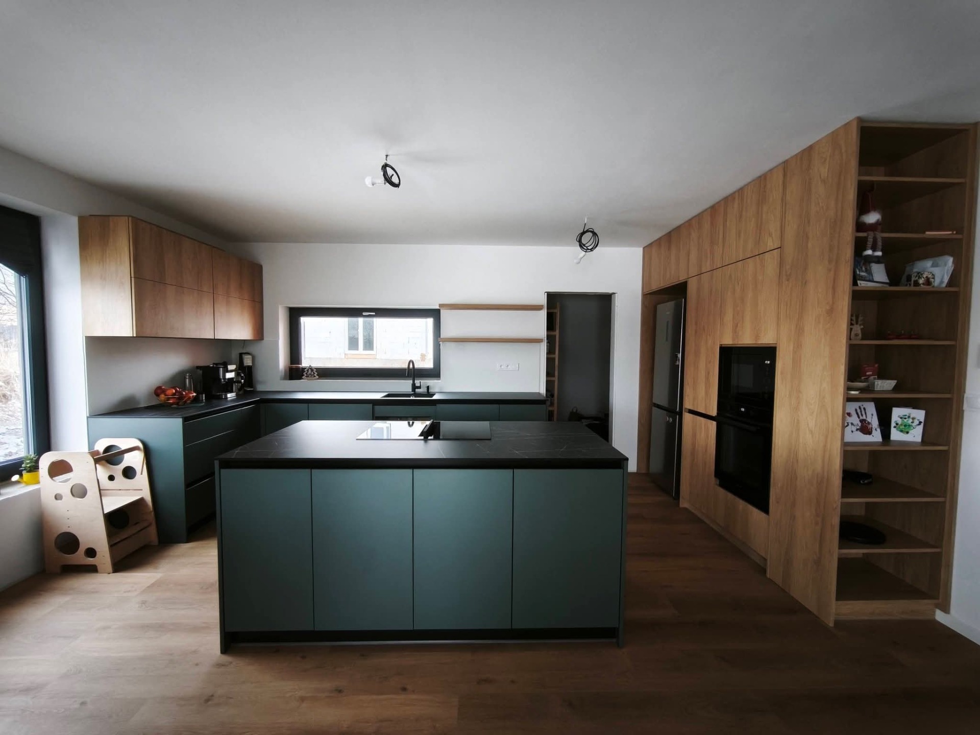Moderní kuchyně s ostrůvkem, která vznikla na míru pro rodinnou novostavbu, spojuje přírodní eleganci s praktickým každodenním využitím. Dominantním prvkem je kombinace dekoru dub Hickory s odstínem kamenné zelené, která prostoru dodává klid, nadčasovost a jemný nádech originality.  Kuchyňský ostrůvek tvoří přirozené centrum dění – místo pro vaření, setkávání i rychlé posezení s rodinou. Celý prostor je navržen s důrazem na maximální komfort a funkčnost, což zajišťují kvalitní výsuvy a výklopné systémy značky Blum. Díky nim je každý centimetr úložného prostoru perfektně využitý a vše máte vždy přehledně po ruce.  Výsledkem je harmonická, moderní kuchyně, která nejen skvěle vypadá, ale především funguje v každodenním životě.