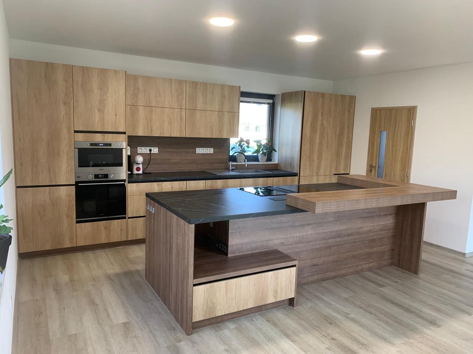  Tahle realizace je ukázkou moderní kuchyně, kde se propojuje přírodní vzhled s funkčností každého detailu.  Dominantou prostoru je kuchyňský ostrůvek, do kterého jsme integrovali varnou desku se spodním odtahem. Díky tomuto řešení zůstává prostor otevřený, vzdušný a ničím nerušený – bez klasické digestoře nad hlavou. Ostrůvek tak přirozeně slouží nejen k vaření, ale i jako místo pro setkávání a každodenní život.  Použitý dekor Dub Nebraska přírodní dodává kuchyni teplo a přirozenost, zatímco pracovní deska Pietra Grigia přináší elegantní kontrast v podobě jemně strukturovaného kamenného vzhledu. Celek působí nadčasově, klidně a zároveň velmi reprezentativně.  Kuchyně je navržená s důrazem na maximální praktičnost – dostatek úložného prostoru, promyšlené rozvržení a kvalitní zpracování zaručují, že bude perfektně sloužit každý den.  Moderní řešení, které spojuje design, komfort a funkčnost v jeden harmonický celek.
