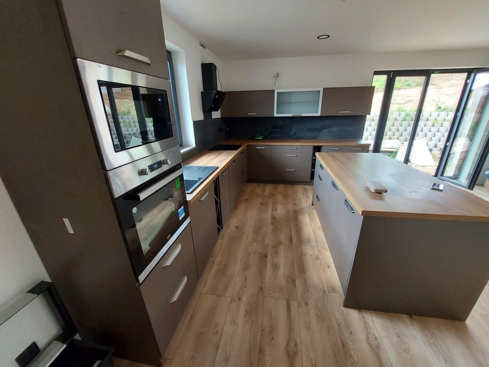 Moderní kuchyň v přírodním duchu 🤎 Kombinace teplého dřevodekoru pracovní desky s elegantními matnými dvířky v hnědo-šedém tónu vytváří nadčasový a útulný prostor. Dominantou je praktický ostrůvek, který nabízí dostatek pracovní plochy i úložného prostoru.  Vestavné spotřebiče, čisté linie a tmavá zástěna dodávají kuchyni moderní charakter, zatímco velká okna krásně propojují interiér se zahradou a přináší spoustu přirozeného světla.  Kuchyň navržená tak, aby byla nejen krásná na pohled, ale hlavně funkční pro každodenní život ✨